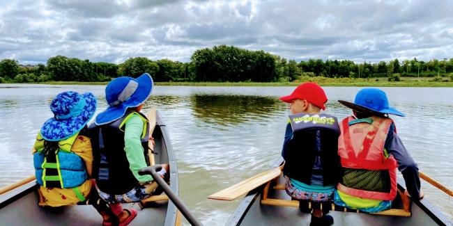 Two pleasure canoes side-by side on the water. Two young children are sitting in each canoe.