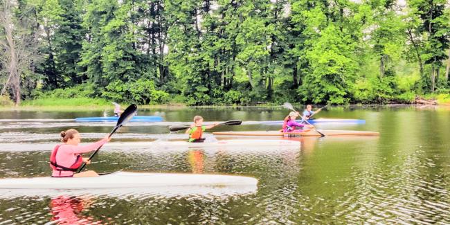 Boys and girls paddle in Sprint kayaks with green trees in the background