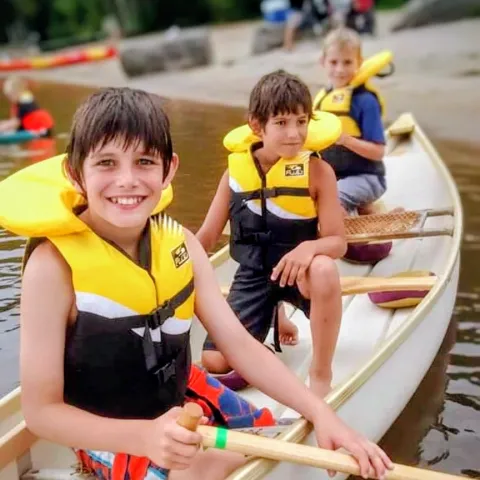 Three boys at the beach, in a white canoe, holding paddles and smiling.